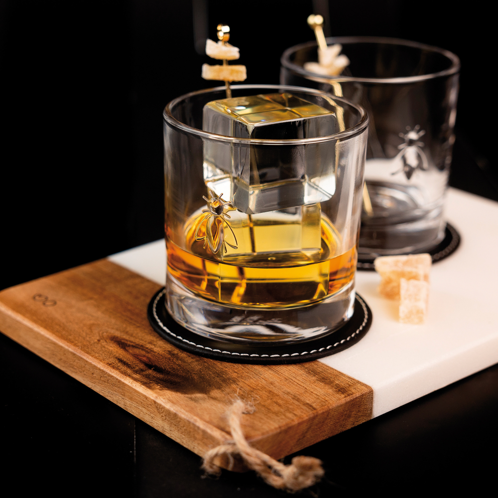 A stylish whiskey glass with a gold bee emblem and a large square ice cube containing amber liquid rests on a two-tone wood and white marble serving board alongside two amber sugar cubes and a matching second glass