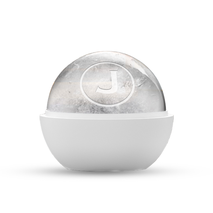 Product photography of a clear ice sphere featuring a prominent white 'J' logo in a circular imprint resting within a smooth white minimalist holder on a black background