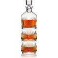 Clear glass whiskey decanter with square stopper stacked on two matching tumblers, all filled with rich amber liquid and featuring elegant fluted and arched patterns reflecting on a white surface