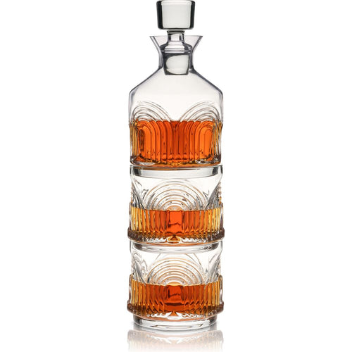 Clear glass whiskey decanter with square stopper stacked on two matching tumblers, all filled with rich amber liquid and featuring elegant fluted and arched patterns reflecting on a white surface