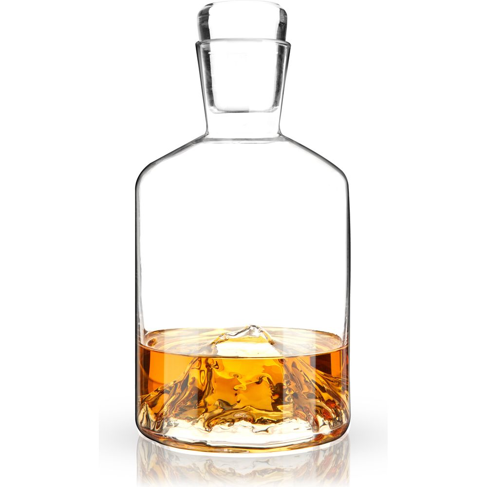 Digital photograph of a unique clear glass whiskey decanter containing golden amber liquid, revealing a dramatic sculpted mountain range design at its base and a matching glass stopper