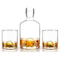 Product photography of a clear glass whiskey decanter and two matching tumblers, each with a unique 3D mountain peak molded into its base, all filled with golden amber liquid on a reflective white surface