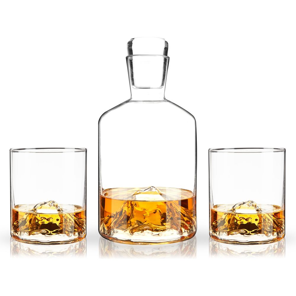 Product photography of a clear glass whiskey decanter and two matching tumblers, each with a unique 3D mountain peak molded into its base, all filled with golden amber liquid on a reflective white surface