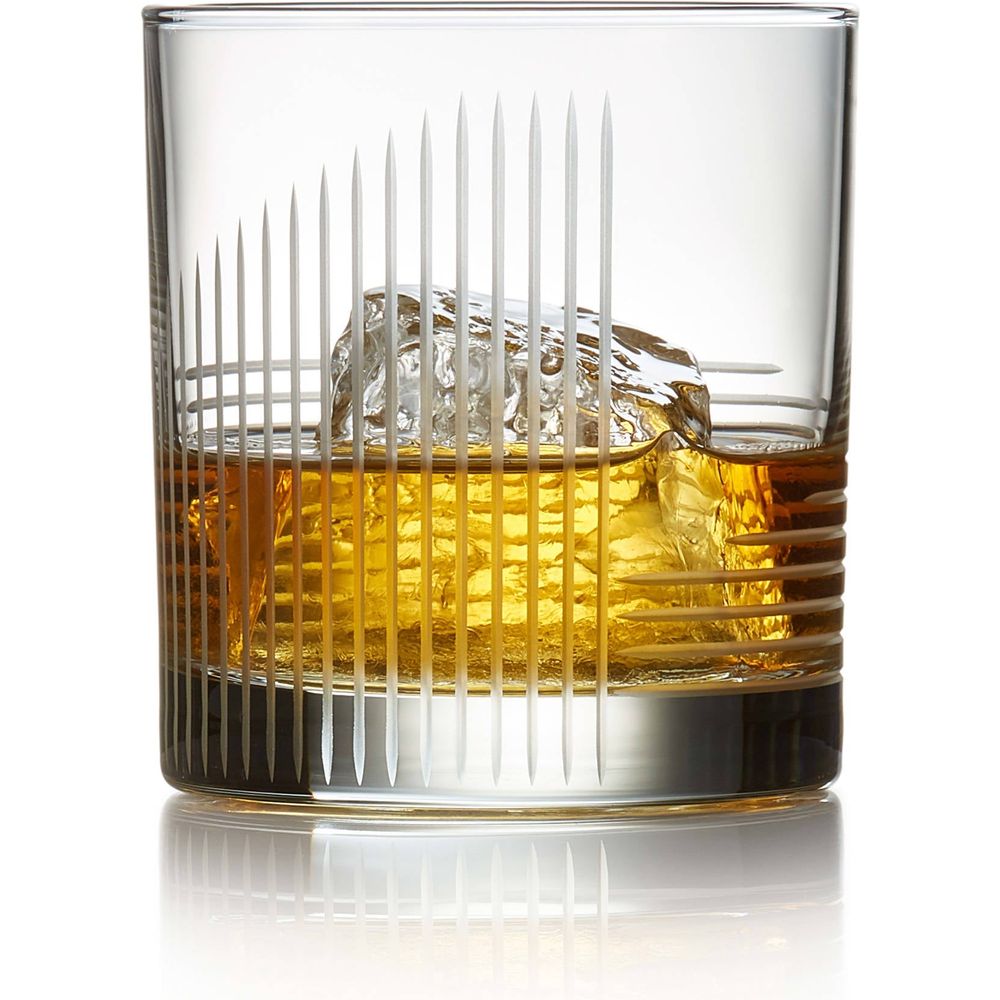 Studio photograph of amber whiskey with a large clear ice cube in an etched glass featuring vertical and horizontal line patterns, reflected on a pristine white surface