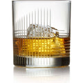 Studio photograph of amber whiskey with a large clear ice cube in an etched glass featuring vertical and horizontal line patterns, reflected on a pristine white surface